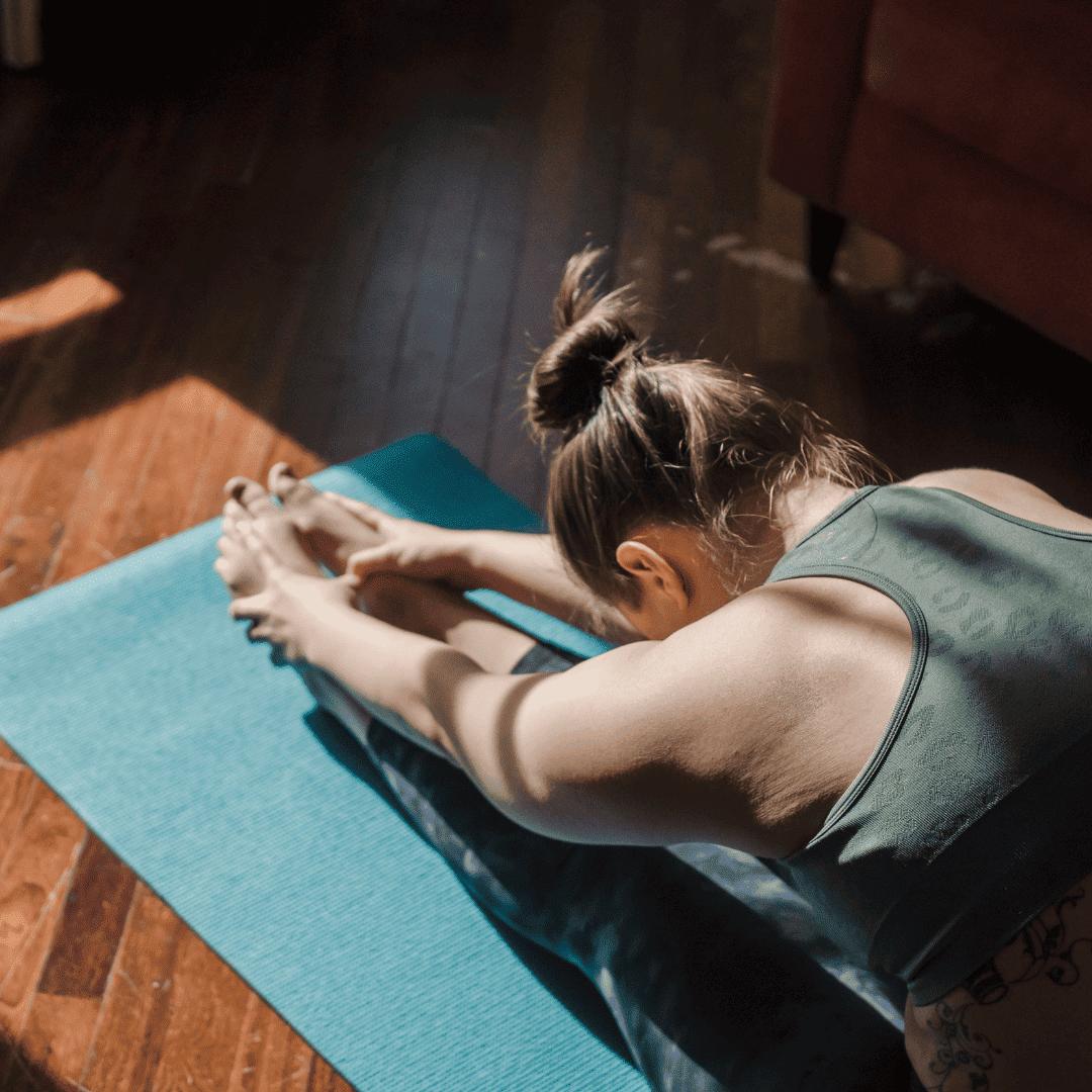 Eine Person in einem grünen Tanktop sitzt auf einer blauen Yogamatte und beugt sich nach vorne, um ihre Zehen zu berühren. Sonnenlicht fällt auf den Holzboden neben ihr.