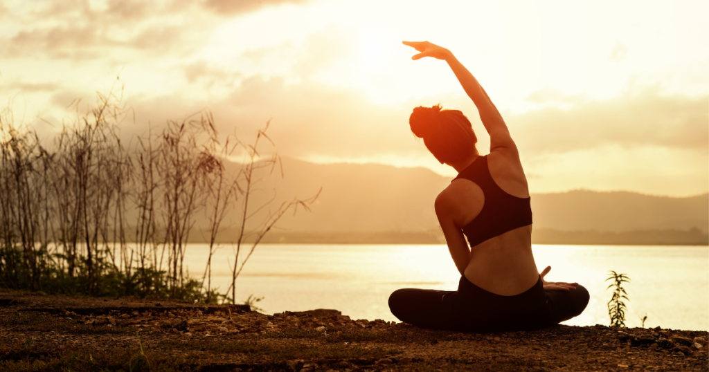 Eine Frau in Sportkleidung sitzt im Schneidersitz am Seeufer und streckt bei Sonnenuntergang einen Arm in den Himmel vor einer Bergkulisse im Licht.