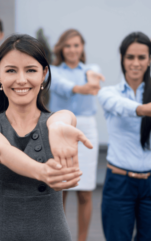 Eine Gruppe von professionell gekleideten Menschen, die im Freien in einer Reihe stehen, lächeln und die Arme nach vorne strecken, während sie an einer Gruppenübung oder einer Teambildungsmaßnahme teilnehmen.